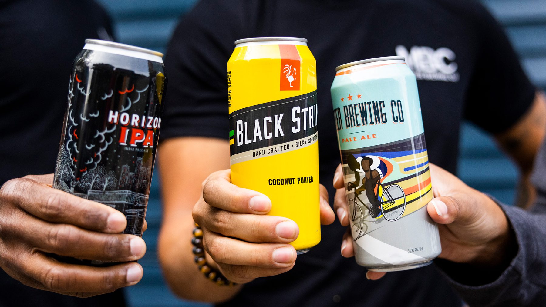 Hands holding cans of Horizon IPA, Black Stripe Coconut Porter, and Metier Brewing Co Pale Ale