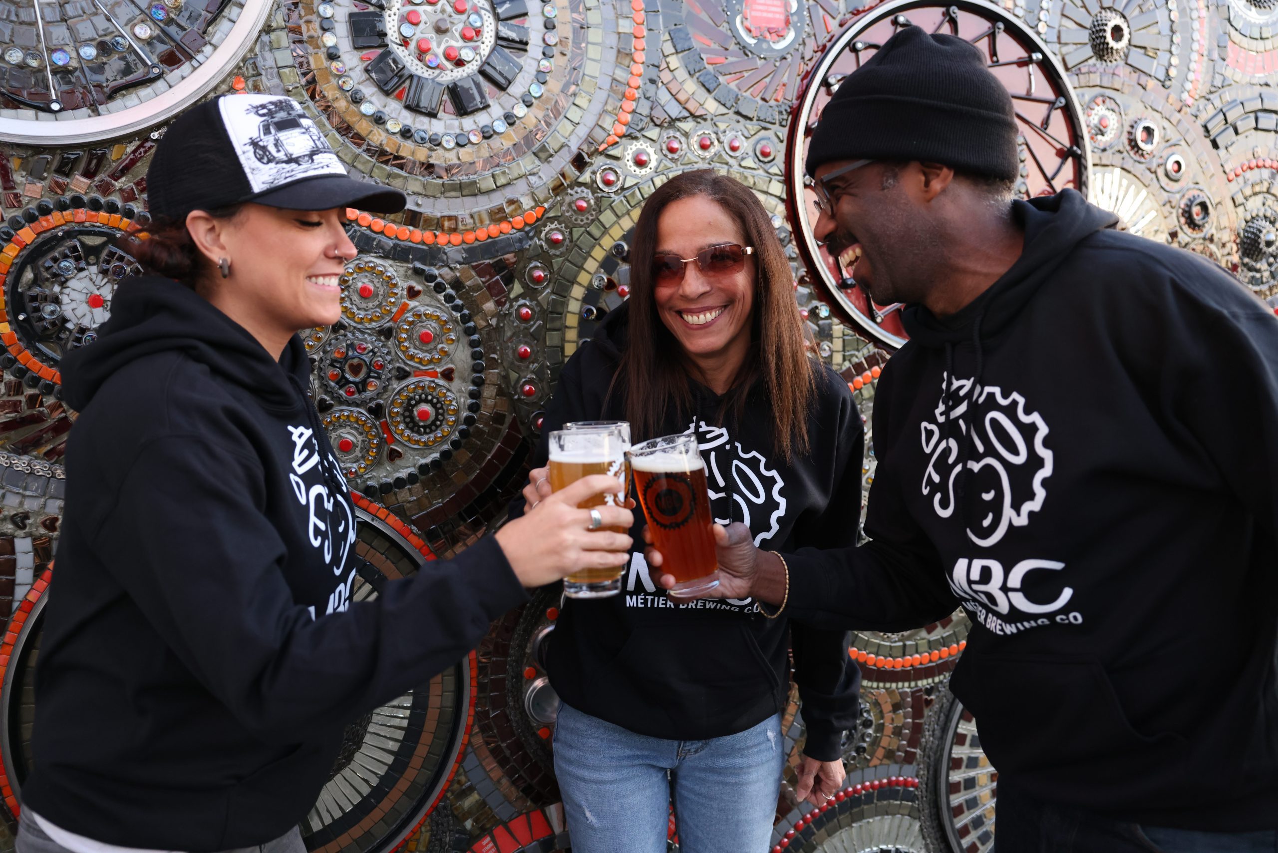 Three people smiling and clinking their beers together all wearing Metier sweatshirts.